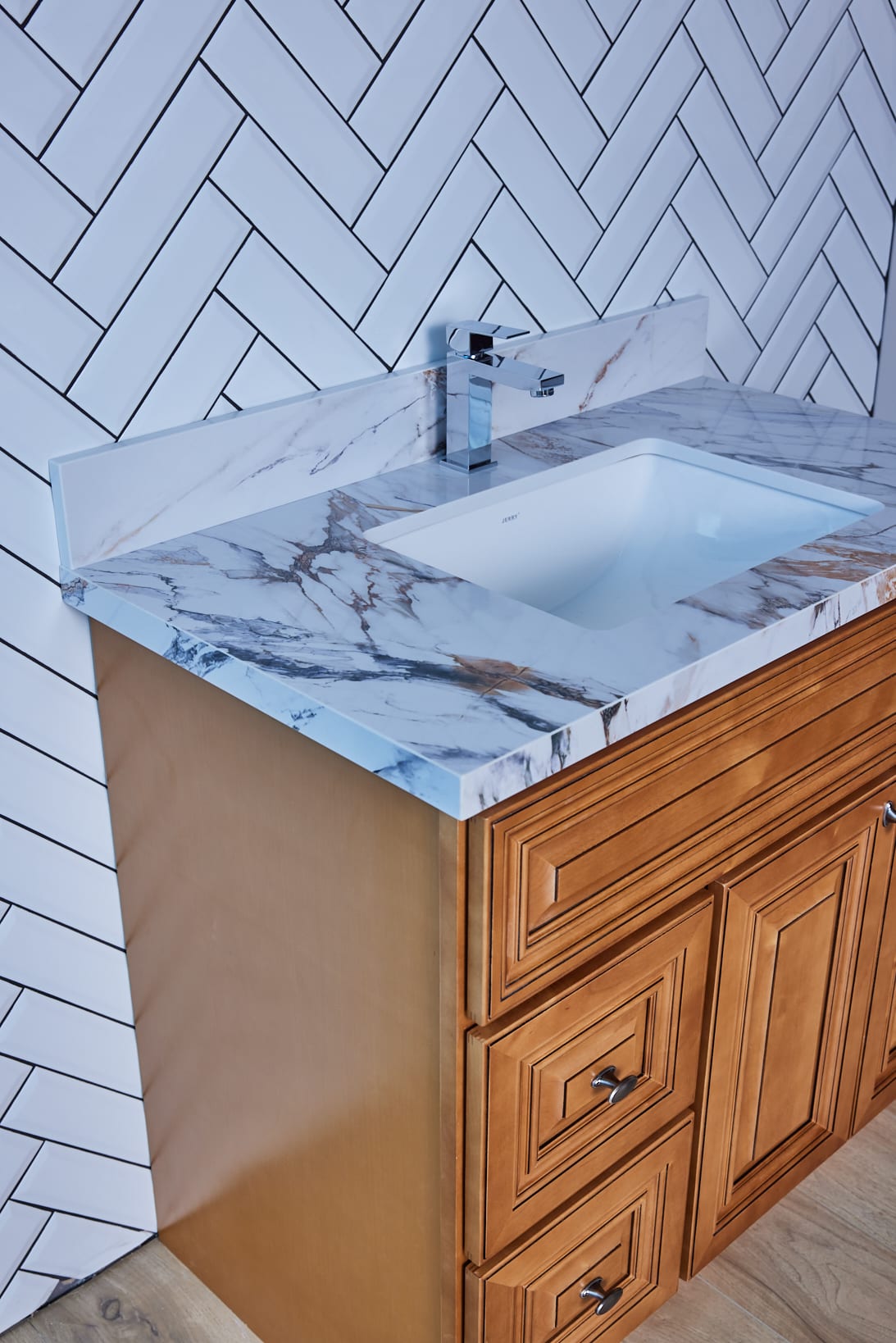 Marble countertop with sink and wooden cabinet against herringbone tile wall.
