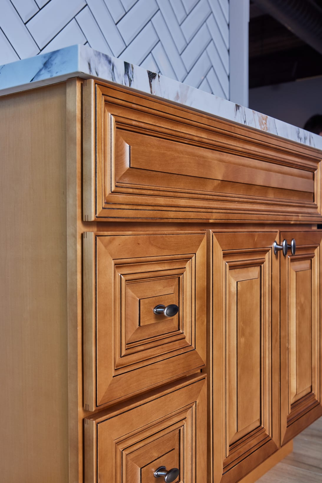 Wooden kitchen cabinet with marble countertop and herringbone tile backsplash.