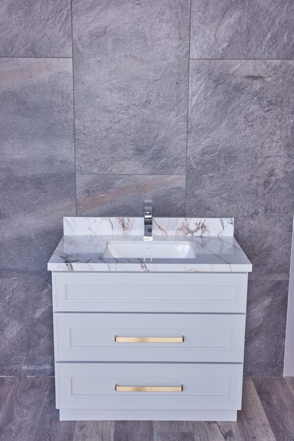 White bathroom vanity with marble countertop and gold handles against gray tile wall.