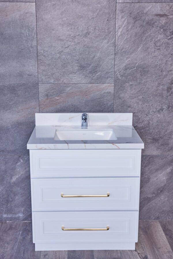 White bathroom vanity with marble countertop and gold handles against gray tile wall.