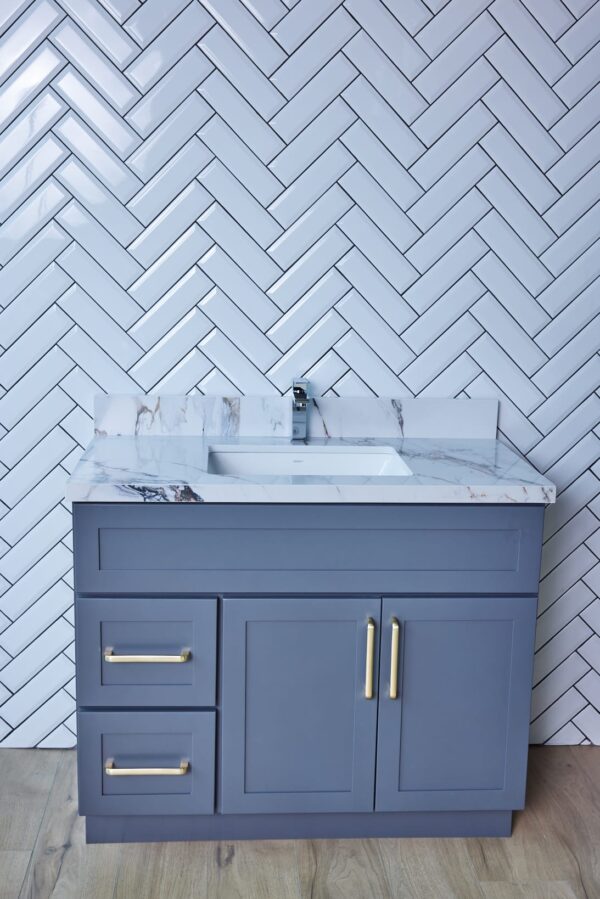 Gray bathroom vanity with marble countertop and herringbone tile backsplash.