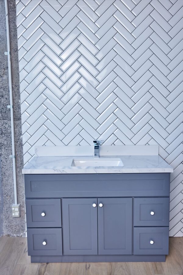Gray bathroom vanity with marble countertop and herringbone tile backsplash.