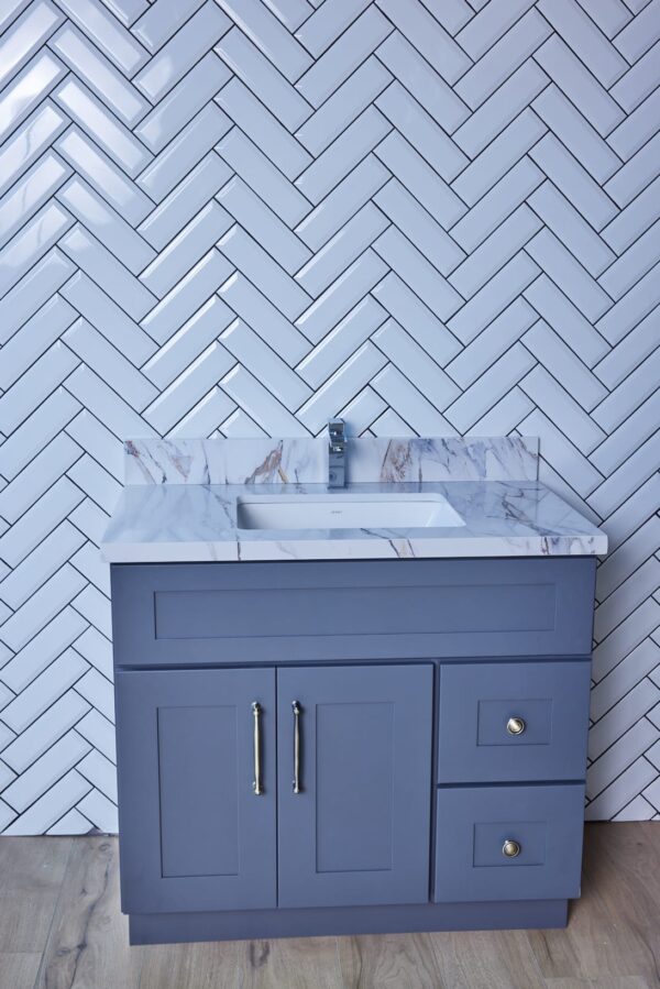 Gray bathroom vanity with marble countertop and herringbone tile backsplash.