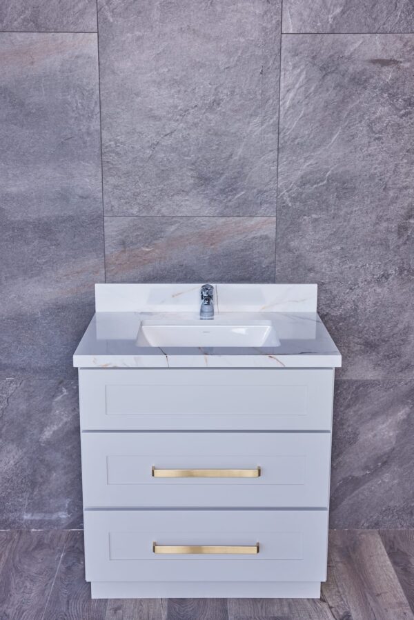White bathroom vanity with marble countertop and gold handles against gray tile wall.