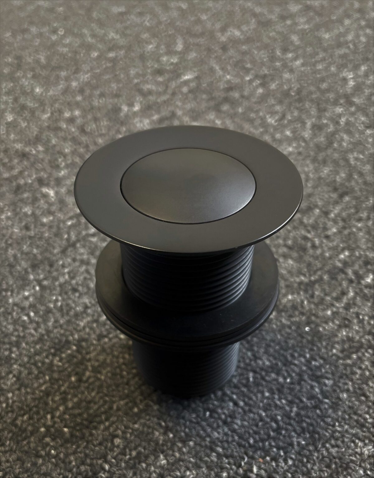 Matte black 32mm pop-up waste on textured surface.