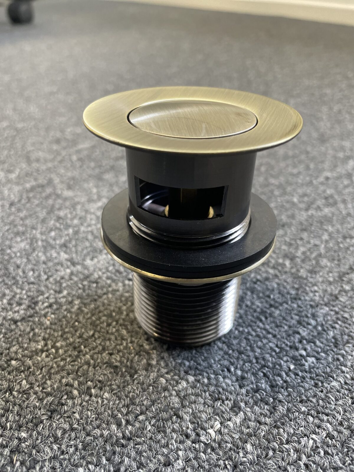 Bronze pop-up waste with overflow on carpeted floor.