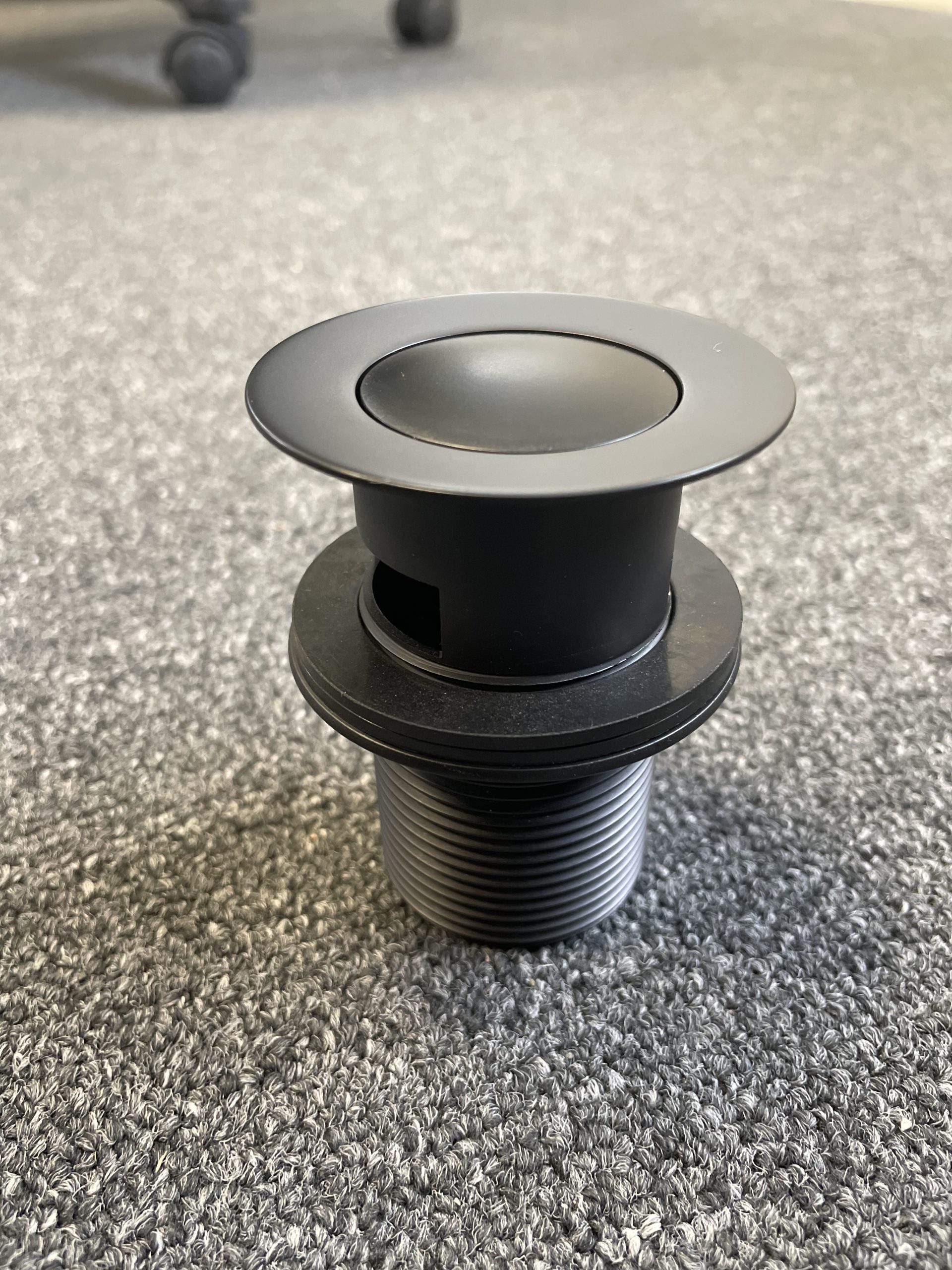 Matt black pop-up waste with overflow on gray surface.