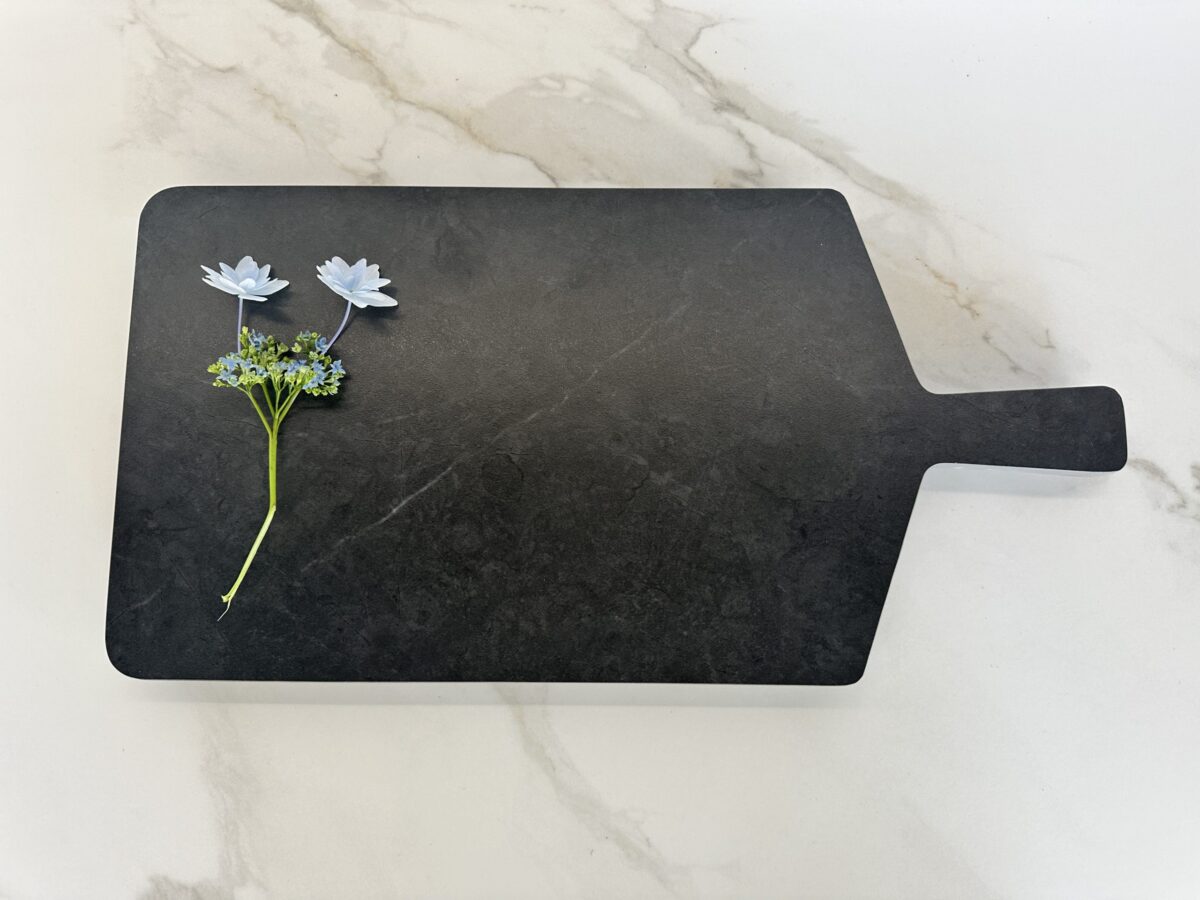 Black cutting board with two white flowers on a marble surface.