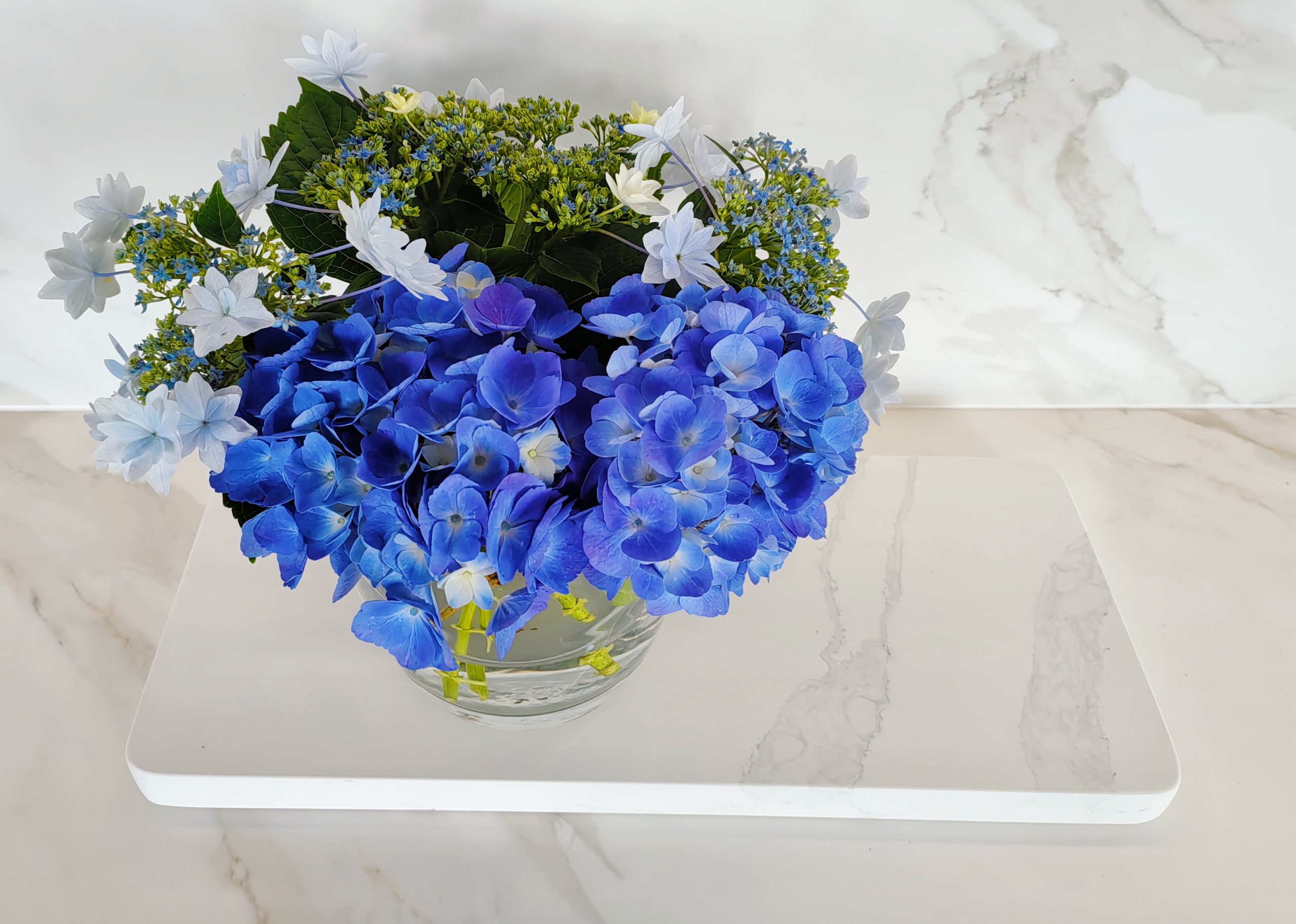 Blue hydrangeas in a glass vase on marble surface.
