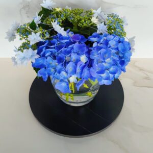 Blue hydrangea and white flowers in a glass vase on a black tray.