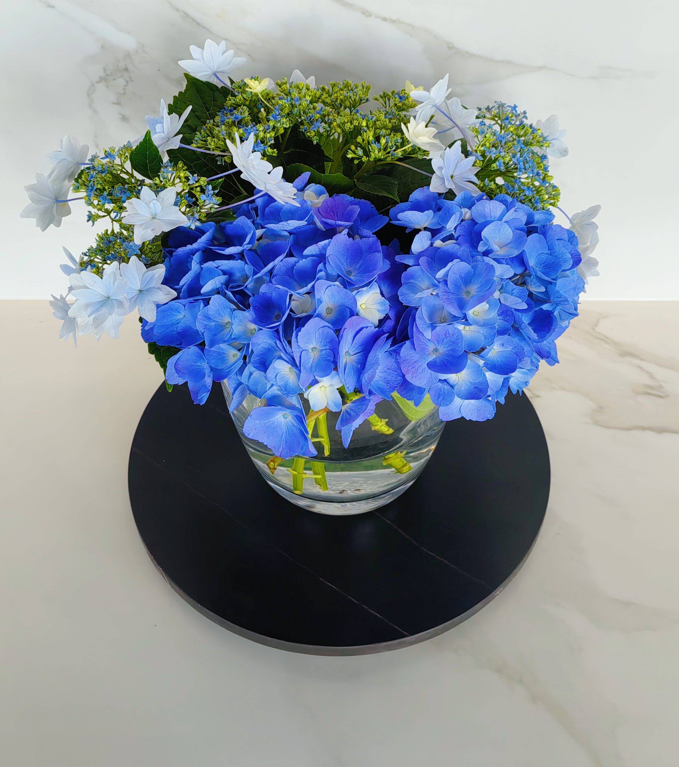 Blue hydrangea and white flowers in a glass vase on a black tray.